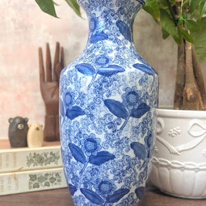 May include: A blue and white ceramic vase with a floral design. The vase has a repeating pattern of blue flowers and leaves on a white background. The vase is on a wooden surface, with plants and other decorative items in the background. The vase is approximately 25 cm tall.