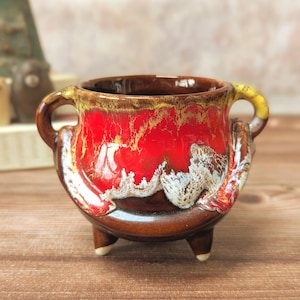May include: A small ceramic bowl with a striking red glaze, featuring yellow and white accents. The bowl has two handles and three small feet. The interior is dark brown. It is likely handmade and suitable for decorative or functional purposes.