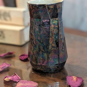Puede incluir: Un jarrón iridiscente facetado con un acabado oscuro y multicolor. Pétalos de rosa secos están esparcidos alrededor del jarrón. El jarrón tiene un diseño geométrico y un interior claro.