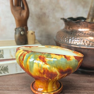 May include: A ceramic pedestal bowl featuring a glossy glaze with swirling patterns of mustard yellow, deep orange, and sage green. The bowl has an oval-shaped rim, a smooth cream-coloured interior, and a circular base that elevates the main body.