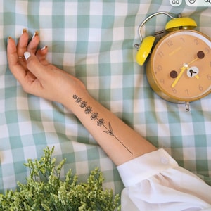 Peut inclure: Un tatouage temporaire à l'encre noire représentant un brin de lavande sur l'avant-bras. Le motif présente une tige verticale avec plusieurs petites grappes de fleurs et deux feuilles à la base. Le tatouage est situé sur l'intérieur du bras.