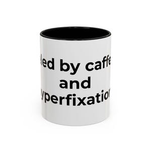 Peut inclure: Mug en céramique blanche avec intérieur noir. Le mug affiche le texte "fueled by caffeine and hyperfixation" en noir, en gras. Un design simple et humoristique pour les amateurs de café.