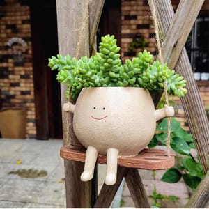 Puede incluir: Una maceta de cerámica beige caprichosa con forma de personaje sonriente, colgada de un columpio de madera con cuerda. La maceta está llena de plantas suculentas verdes. El diseño incluye brazos, piernas y una cara feliz.