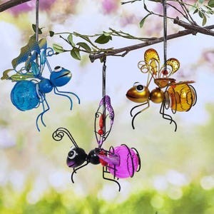 May include: Three hanging metal insect ornaments: a blue ant, a gold bee, and a purple and black ant. The insects have wire bodies and colorful glass orbs for their abdomens. They are suspended from a tree branch against a blurred background.