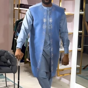 May include: A light blue formal suit with silver embellishments. The outfit includes a long vest, a long-sleeved shirt, and matching pants. The person is wearing a matching cap and black shoes. A walking stick is held in the right hand.
