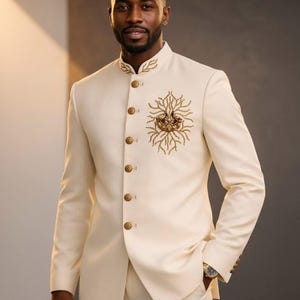 May include: Ivory suit with a mandarin collar and gold-toned buttons. The jacket features an embroidered floral design on the chest. The suit is tailored with a slim fit and is suitable for formal occasions. The model is wearing a watch.