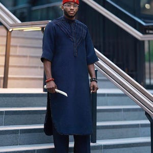 May include: A navy blue traditional outfit consisting of a long tunic and matching pants. The tunic has decorative stitching and is paired with a red hat with a white feather. The outfit is accessorized with bracelets.