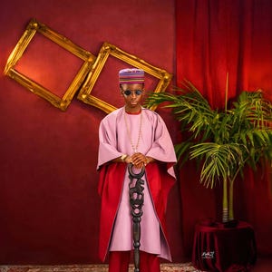 May include: A person wearing a pink and red traditional outfit, a patterned hat, and sunglasses, holding a carved wooden staff. The background features gold picture frames, red curtains, and a potted plant. The outfit includes a long necklace and black shoes.