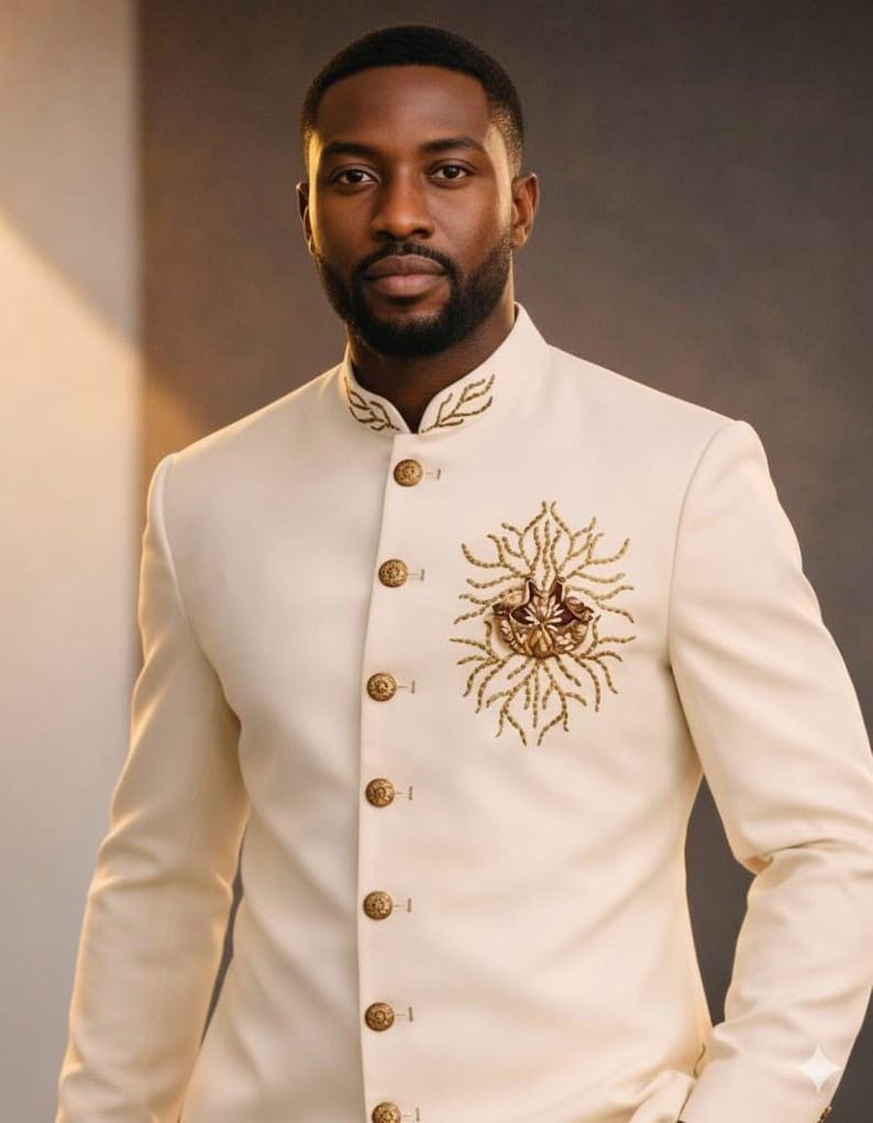 May include: Ivory suit jacket with a mandarin collar and gold-colored buttons. The jacket features intricate gold embroidery on the chest and collar, creating a formal and elegant look. The suit is tailored with a classic fit.