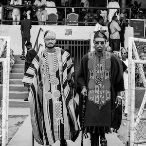 May include: Two men in traditional African attire walk side-by-side. One wears a striped robe with a matching head covering and beaded necklaces. The other is in a dark robe with a patterned design, sunglasses, and a walking stick. The image is in black and white.