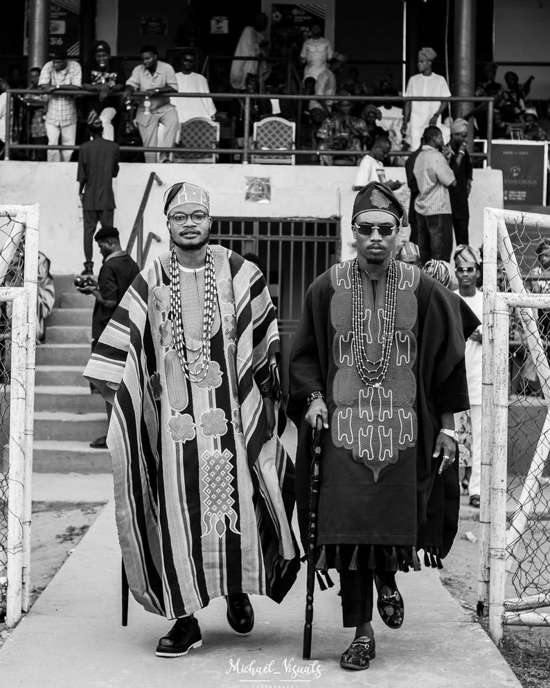 May include: Two men in traditional African attire walk side-by-side. One wears a striped robe with a matching head covering and beaded necklaces. The other is in a dark robe with a patterned design, sunglasses, and a walking stick. The image is in black and white.