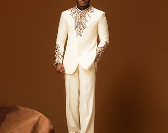 May include: Ivory suit with intricate gold and brown embroidery on the collar and sleeves. The suit features a tailored jacket and wide-leg pants. The person is wearing brown leather shoes. The background is a solid brown.