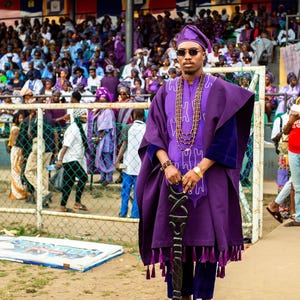 May include: A person in a purple traditional outfit, including a hat, tunic, and pants, stands holding a decorative staff. The outfit is adorned with tassels and a beaded necklace. The background shows a crowd of people, many also wearing purple.