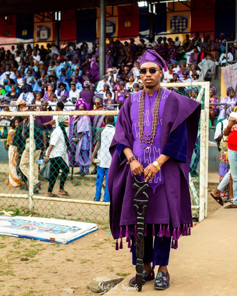 May include: A person in a purple traditional outfit, including a hat, tunic, and pants, stands holding a decorative staff. The outfit is adorned with tassels and a beaded necklace. The background shows a crowd of people, many also wearing purple.