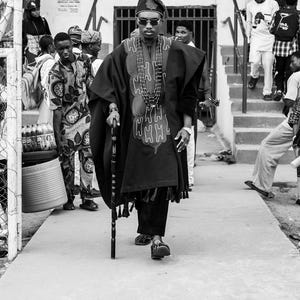 May include: A black and white photograph of a person wearing traditional African attire, including a robe with an embroidered design, a hat, and beaded necklaces. The person is holding a cane and walking on a paved surface.