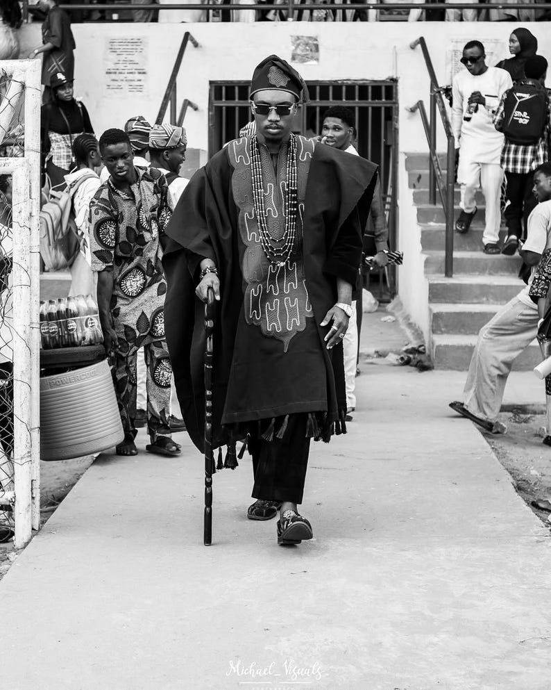May include: A black and white photograph of a person wearing traditional African attire, including a robe with an embroidered design, a hat, and beaded necklaces. The person is holding a cane and walking on a paved surface.
