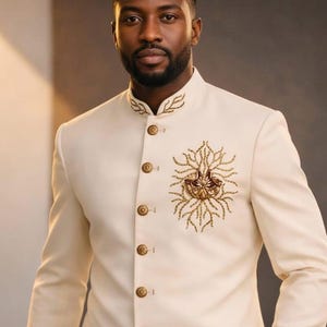 May include: Ivory-colored suit jacket with a mandarin collar and gold-colored buttons. The jacket features intricate gold embroidery on the chest and collar, creating a formal and elegant look. The suit is tailored with a classic fit.