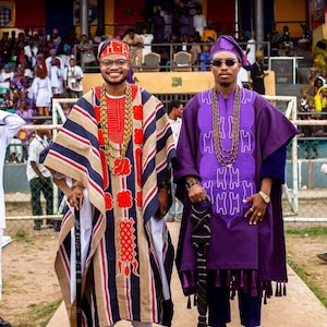 May include: Two people wearing traditional African attire. One person is in a striped robe with red and gold accents, a matching head covering, and a walking stick. The other person is in a purple robe with a matching head covering and a walking stick.