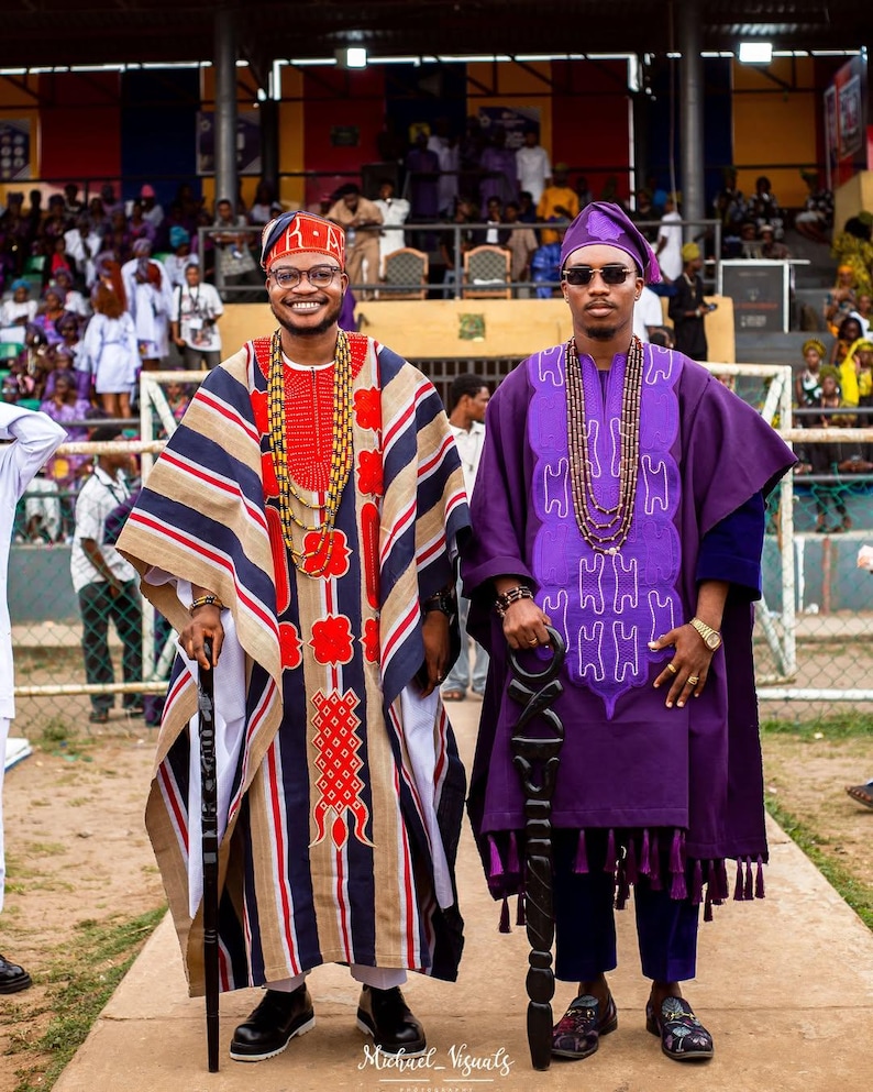May include: Two people wearing traditional African attire. One person is in a striped robe with red and gold accents, a matching head covering, and a walking stick. The other person is in a purple robe with a matching head covering and a walking stick.