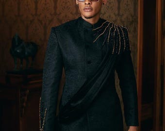 May include: A black suit with a textured pattern, featuring gold beaded accents on the shoulder and sleeve. The suit includes a draped sash and is paired with sunglasses and a decorative hat.