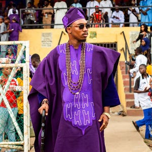 May include: A person wearing a purple traditional outfit, including a hat and robe, with intricate white embroidery. The outfit is accessorized with a beaded necklace, sunglasses, and a walking stick. The image is taken outdoors.