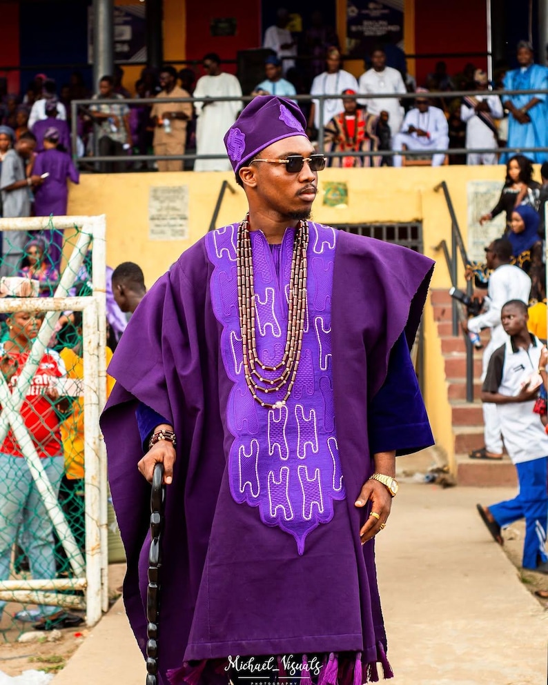 May include: A person wearing a purple traditional outfit, including a hat and robe, with intricate white embroidery. The outfit is accessorized with a beaded necklace, sunglasses, and a walking stick. The image is taken outdoors.