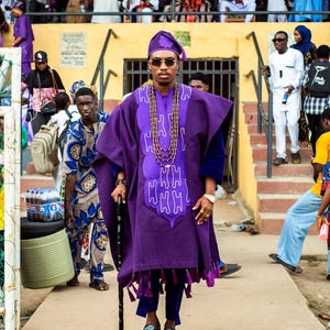 May include: A person in a purple traditional outfit, including a hat, tunic, and pants, stands with a cane. The tunic has a decorative design and fringe. The person wears sunglasses and a beaded necklace. Other people are in the background.