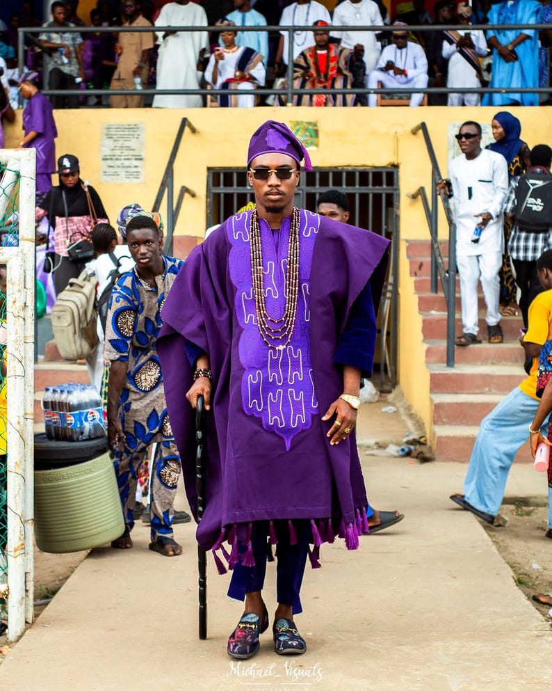 May include: A person in a purple traditional outfit, including a hat, tunic, and pants, stands with a cane. The tunic has a decorative design and fringe. The person wears sunglasses and a beaded necklace. Other people are in the background.