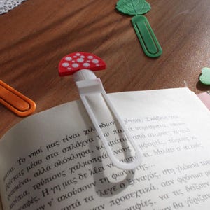 May include: Four plastic bookmarks featuring distinct shapes: a red and white mushroom, an orange maple leaf, a green leaf, and a light green four-leaf clover. One mushroom bookmark is clipped onto the top of an open book with printed Greek text.