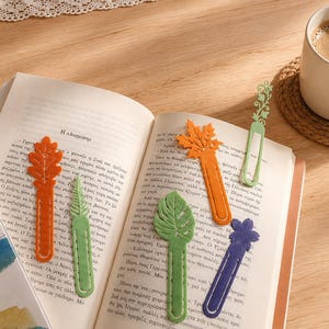 May include: A collection of five leaf-shaped bookmarks in orange, light green, dark green, and purple, resting on an open book. The bookmarks feature intricate cut-out designs and stitching details, with one bookmark displaying a vine-like pattern.