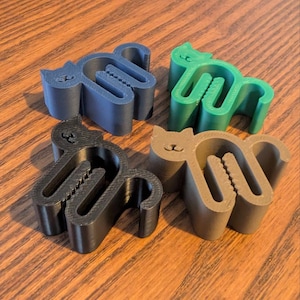 May include: Four cat-shaped cable tidies in dark blue, green, black, and brown. Each organiser features a stylised feline silhouette with a central slotted opening designed to hold and secure cables, with a serrated edge along the inner channel.