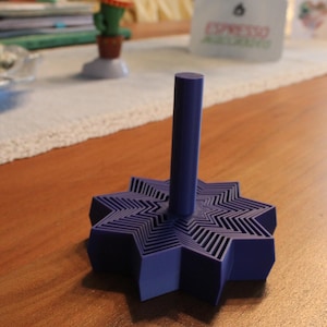 May include: A dark purple spinning top featuring a geometric star-shaped base with a layered, concentric design and a smooth, cylindrical vertical handle extending from the centre, crafted with a matte finish and a solid, uniform colour throughout.