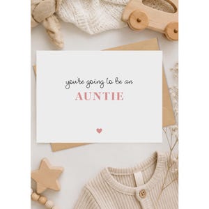 May include: A white rectangular greeting card featuring the black text you're going to be an and the pink text AUNTIE, with a small pink heart icon centered below the text. The card is displayed on a neutral surface with various baby items.