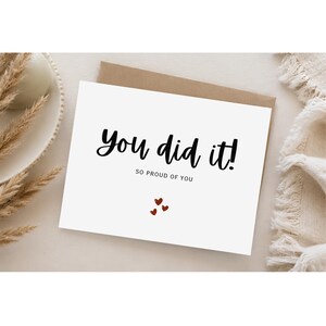 Może przedstawiać: Biała kartka okolicznościowa z brązową kopertą, zawierająca czarny napis You did it! oraz mniejszy czarny napis SO PROUD OF YOU, uzupełniona trzema małymi czerwonymi sercami ułożonymi w grupę pod tekstem na przedniej stronie kartki.