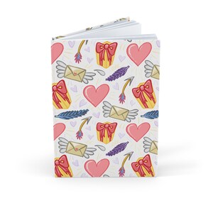 May include: A white journal with a repeating pattern of pink hearts, winged envelopes, gift boxes with red bows, arrows, and purple feathers. The journal is open, revealing blank pages.