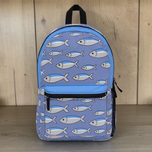 Puede incluir: Una mochila azul con un patrón repetido de peces blancos y azules. La mochila tiene una cremallera negra, un bolsillo delantero azul y un asa negra. El fondo es de madera clara.