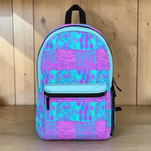 May include: A colourful backpack with a geometric pattern. The backpack has a light blue top and front pocket, with a black zip and handle. The main body of the backpack has a patchwork design in shades of purple, pink, and teal.