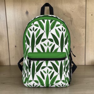 May include: A white backpack with a green leaf pattern. The backpack has a green front pocket and trim, black straps, and a black zipper. The bag is made of a durable material and is suitable for carrying books and other items.