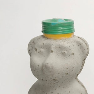 May include: Two gray concrete bear-shaped bottles with green and blue plastic lids. The bottles are sitting on a gray surface.