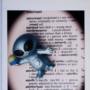 Puede incluir: Una figurita de extraterrestre azul claro con ojos grandes y una expresión traviesa. La figurita sostiene un objeto pequeño y redondo. El fondo es una página de diccionario con la definición de la palabra "miscreant".
