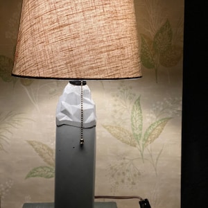 Może przedstawiać: Szara betonowa lampa z geometrycznym wzorem i brązowym abażurem z tkaniny. Lampa stoi na ciemnoszarym podłożu.