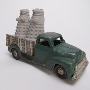 Peut inclure: Un camion jouet vert vintage à l'aspect usé. La benne du camion transporte deux figurines grises en forme de cône avec une texture cloutée. Le camion a des accents argentés sur le pare-chocs avant et les roues.