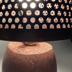 May include: A modern table lamp with a dark metal shade featuring a pattern of circular cutouts. The base is a reddish-brown, textured stone. The lamp is lit, with the light shining through the holes in the shade.