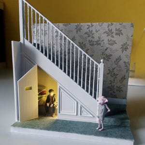 Puede incluir: Un diorama en miniatura que presenta una escalera blanca con un pequeño armario debajo. Dentro del armario iluminado se sienta una pequeña figura. Una criatura rosa se encuentra en el suelo verde junto a la escalera, frente a un papel tapiz floral.