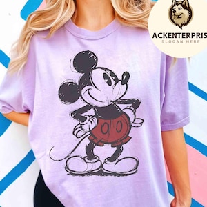 Puede incluir: Una camiseta de color lila claro con una imagen esbozada de Mickey Mouse en negro, rojo y blanco. La camiseta tiene mangas cortas y cuello redondo. La imagen incluye el texto "ACKENTERPRISE. SLOGAN HERE" en un logotipo circular.