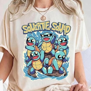 以下が含まれることがあります： アイボリーのTシャツ。黄色い甲羅を持つ5匹の青いカメのキャラクターが描かれたグラフィックプリントで、1匹は黒いサングラスをかけ、青い水しぶきに囲まれています。キャラクターの上には「SQUIRTLE SQUAD」という文字が黄色でプリントされています。