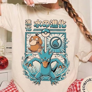 May include: An ivory t-shirt featuring a graphic print of a brown Psyduck and a blue Golduck with water effects. The design includes a Poké Ball icon, a water droplet symbol, and Japanese text that reads "水の進化" above the characters.