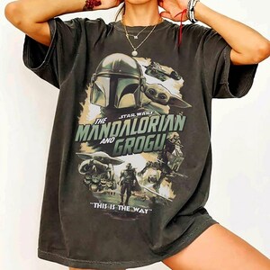 K&ouml;nnte beinhalten: Dunkelgraues T-Shirt mit einem Grafikdesign aus der Star Wars-Serie, "The Mandalorian and Grogu." Das Design zeigt den Helm des Mandalorianers, Grogu und den Schriftzug "THIS IS THE WAY." Das T-Shirt hat einen Vintage-Look.
