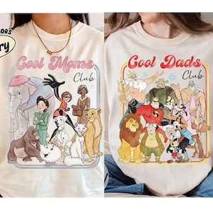 Puede incluir: Dos camisetas color crema con el texto "Cool Moms Club" y "Cool Dads Club" en un diseño retro. La camiseta "Cool Moms" presenta personajes de dibujos animados, y la camiseta "Cool Dads" tiene un conjunto diferente de personajes.