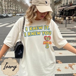 K&ouml;nnte beinhalten: Ein elfenbeinfarbenes T-Shirt mit dem blau-gelben Text I KNEW IT, I KNEW YOU, einem roten Herz mit den Buchstaben TS und dem Wort STORY &uuml;ber der gelben Zahl 5. Das T-Shirt wird mit einer schwarzen Umh&auml;ngetasche und einer beigen Baseballkappe kombiniert.
